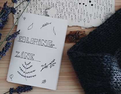 Picture with a folded white paper booklet with 'coloring zine' on the cover